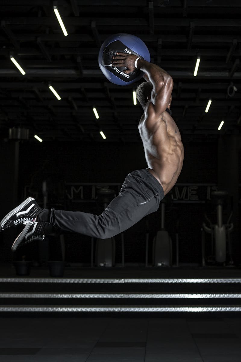 Muscular athlete performing a high jump with a medicine ball in a gym, showcasing strength and fitness.